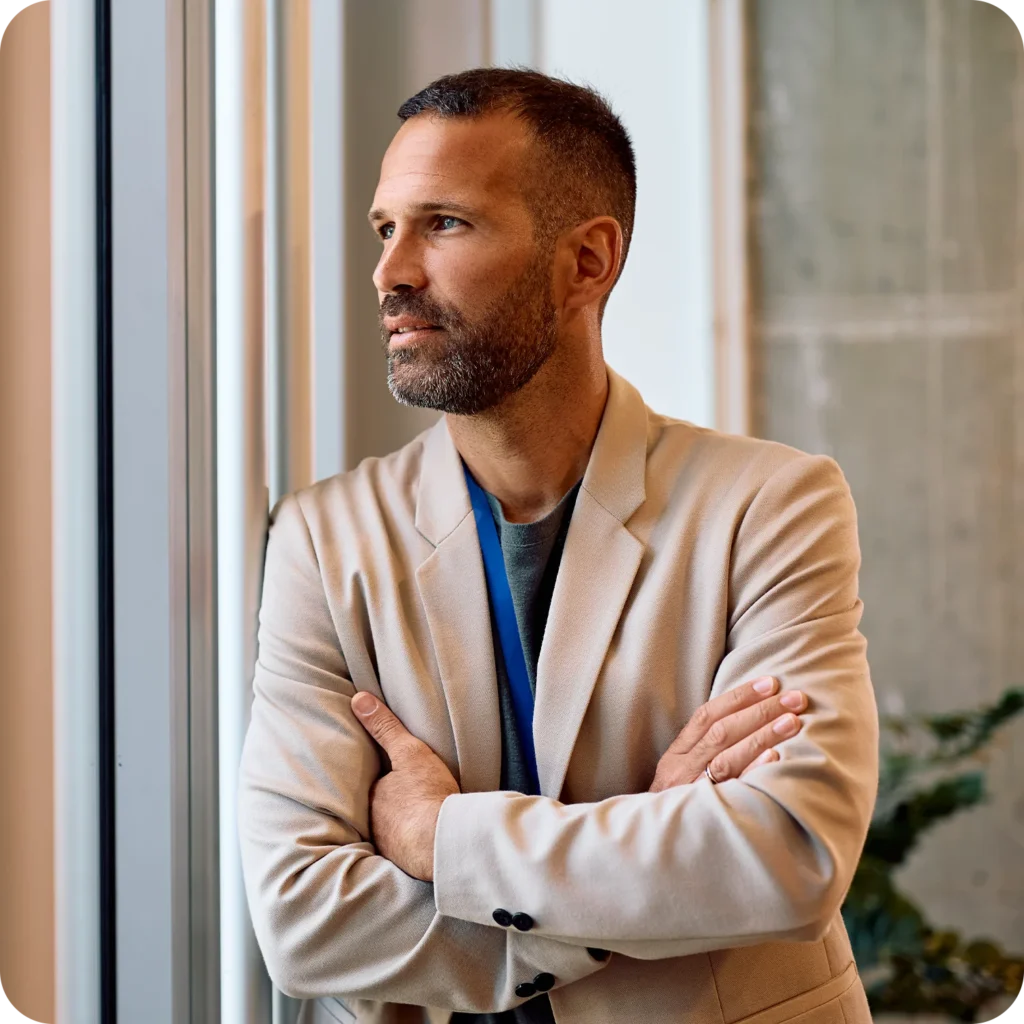 Businessman standing with folded arms by an office window, looking thoughtful.