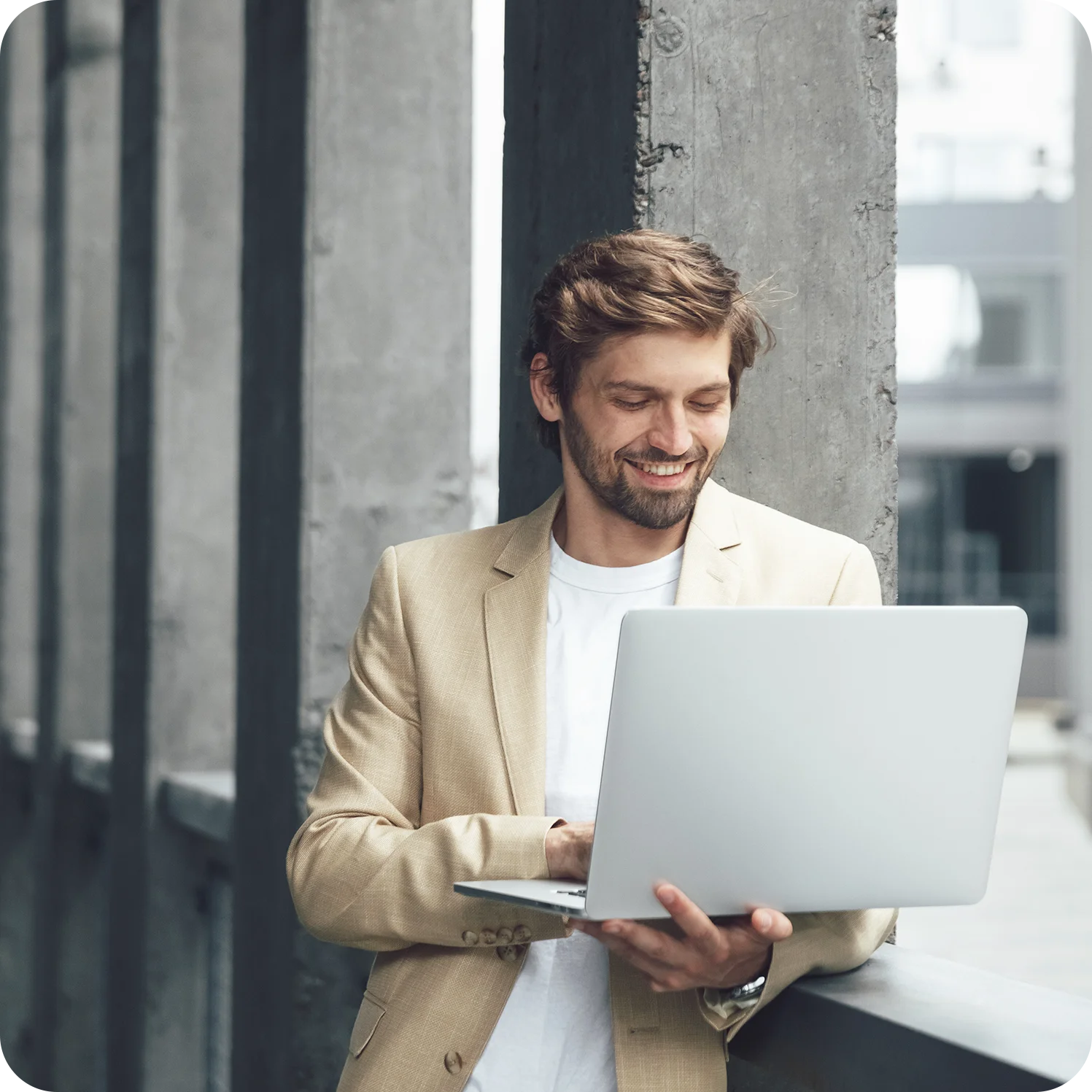 Smiling businessman in a beige blazer using a laptop in a modern office setting.