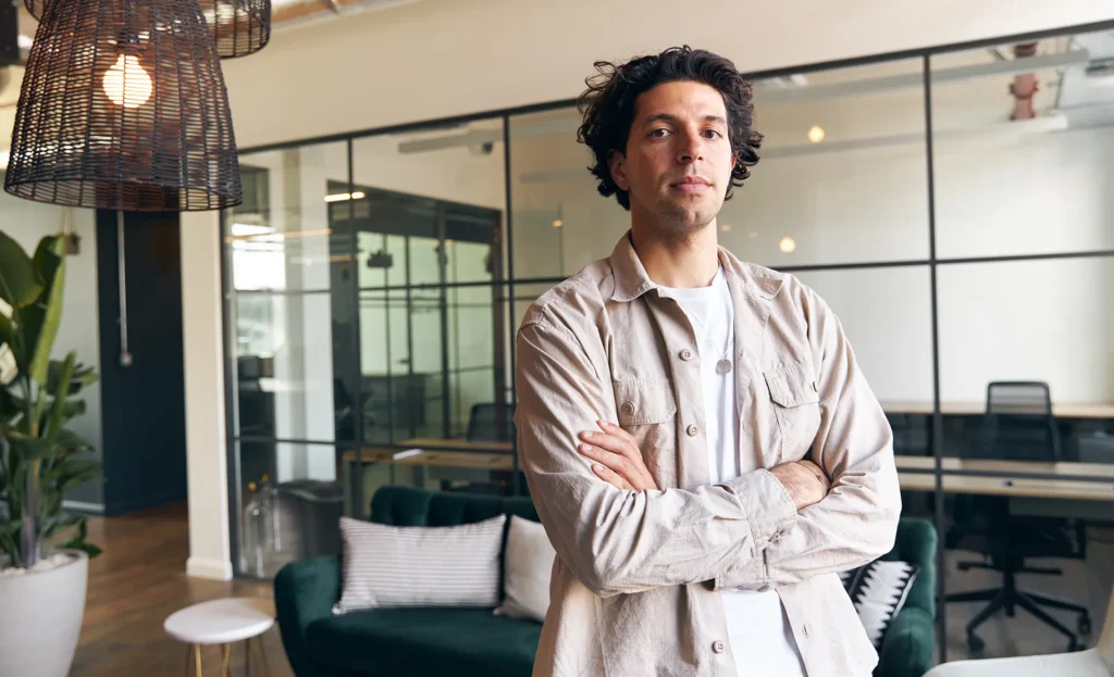 A confident man with curly dark hair stands with arms crossed in a modern, open-plan office.